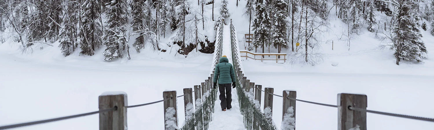 The image depicts a tranquil winter scene where an individual is walking on a wooden bridge covered in snow. The bridge, which has wooden railings and is supported by cables, extends into the distance and is surrounded by a snowy landscape.