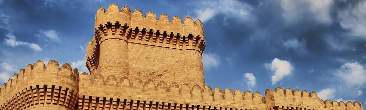 The image shows a panoramic view of an ancient castle made of brown stone. The castle has crenellated walls and towers, indicative of medieval architecture. 