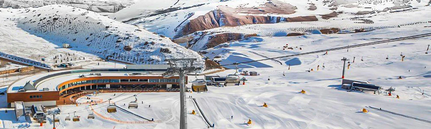 he image captures a wide, panoramic view of a snowy mountainous landscape. A modern ski resort with an orange and white exterior is prominently featured in the middle of the image. 