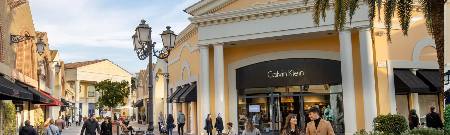The image captures a bustling shopping center with various stores and a crowd of people. A prominent Calvin Klein store is in the foreground with its name displayed in large white letters against a black background. 
