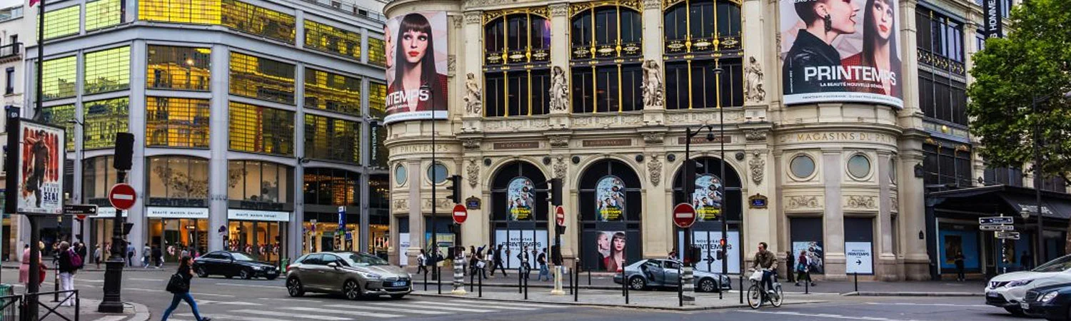 The image captures a bustling street scene in front of the Printemps department store in Paris. The Printemps building is architecturally rich, featuring a mix of modern and classical elements, with large windows and intricate detailing.